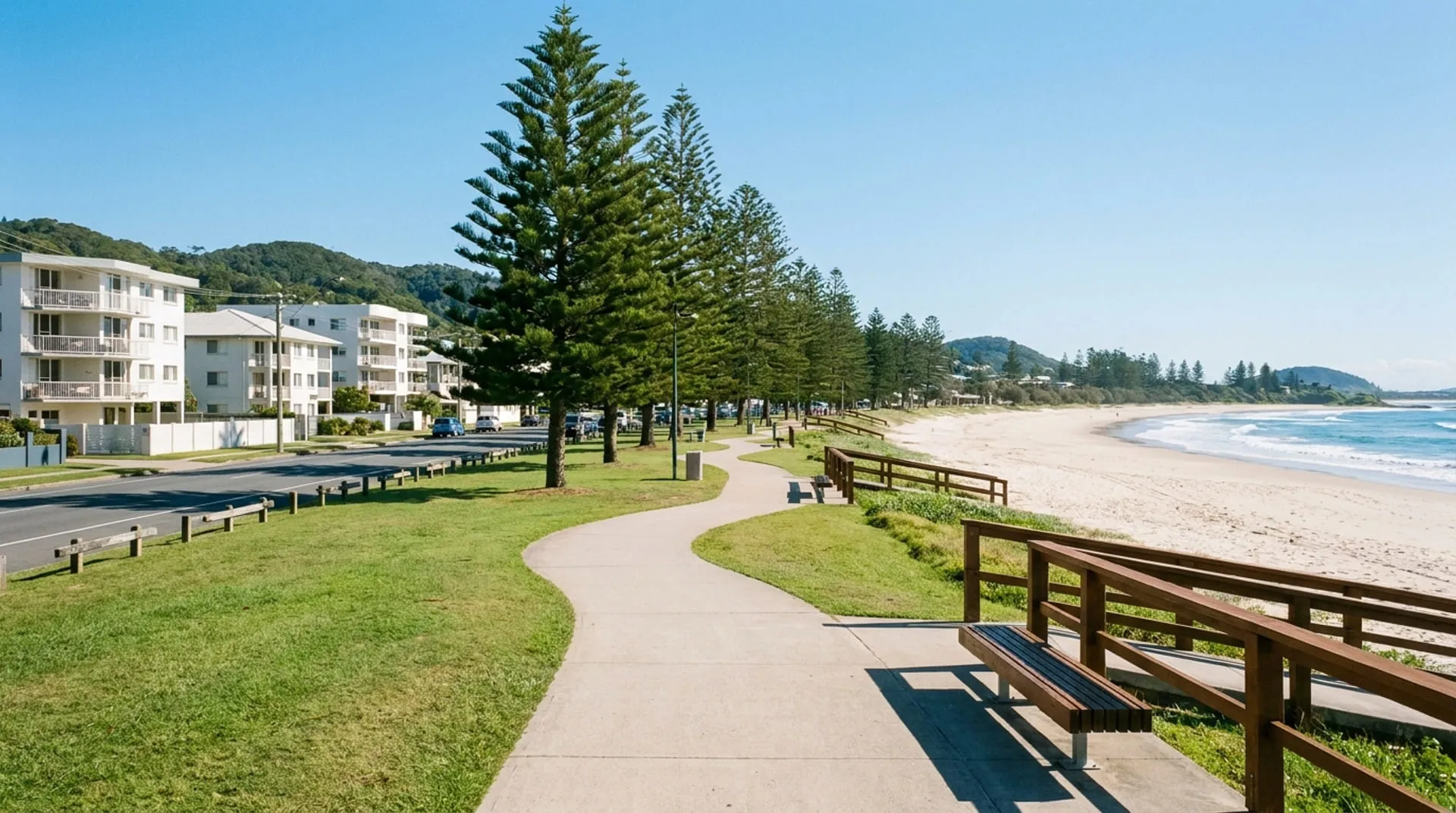A beachfront promenade beside a grassy reserve of Norfolk pines above a wide beach