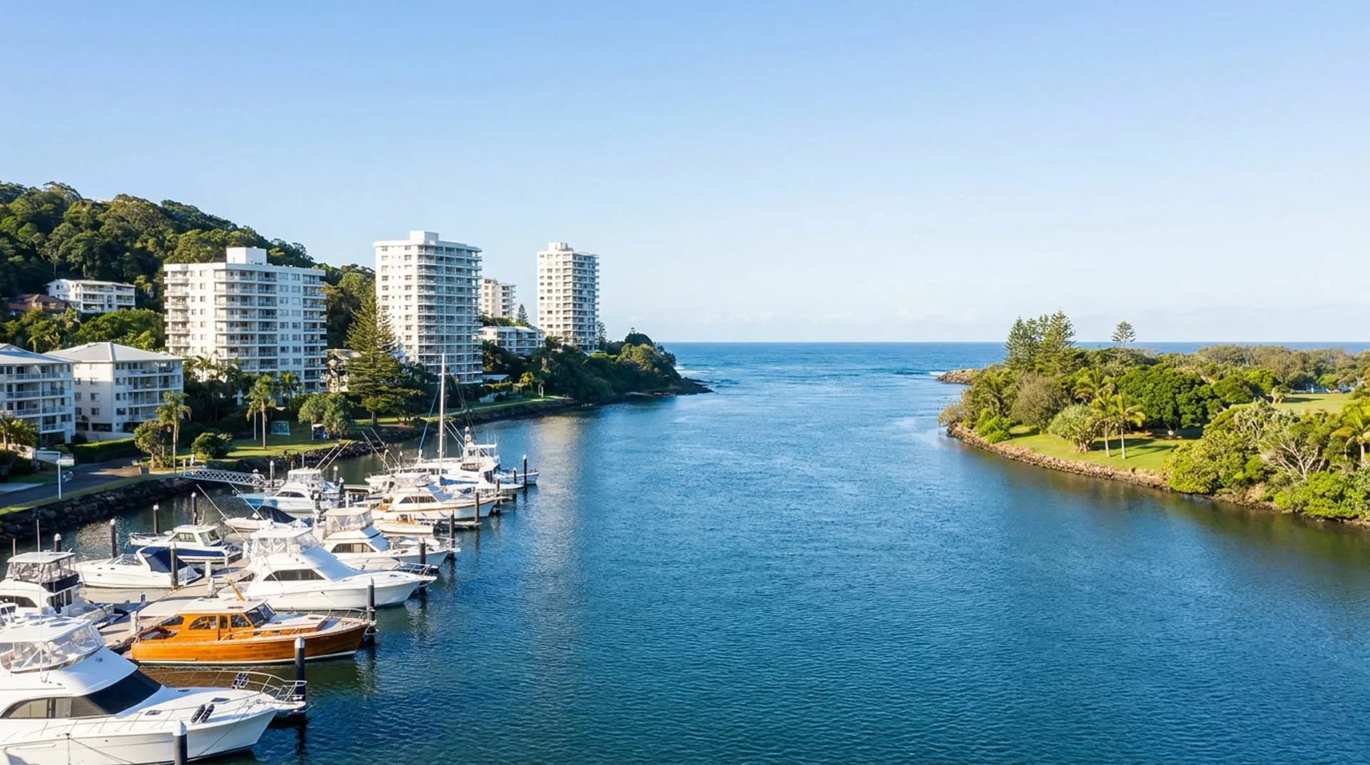 A marina on a broad river mouth with mid-rise apartment towers and a headland beyond
