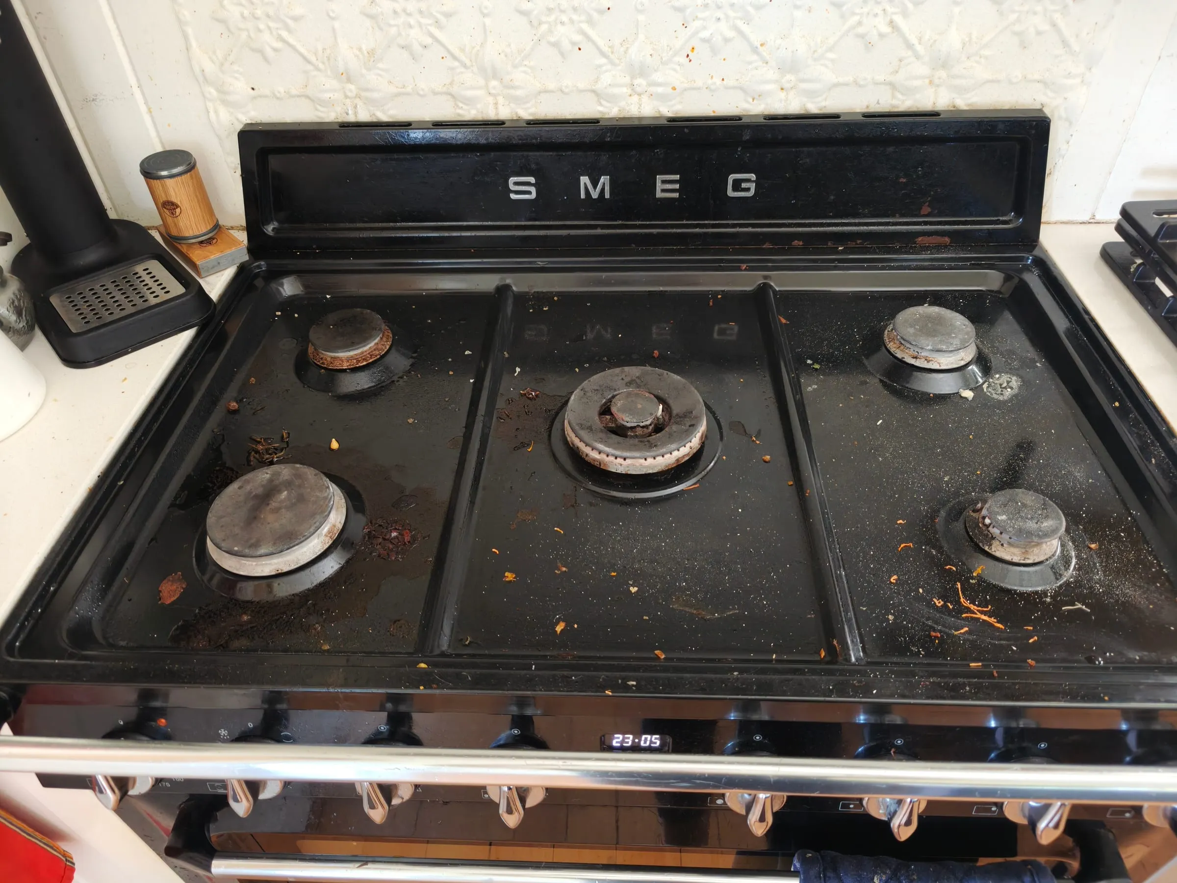 A gas cooktop before a TLB deep clean, with spilled coffee grounds, crumbs and food scraps across the black enamel
