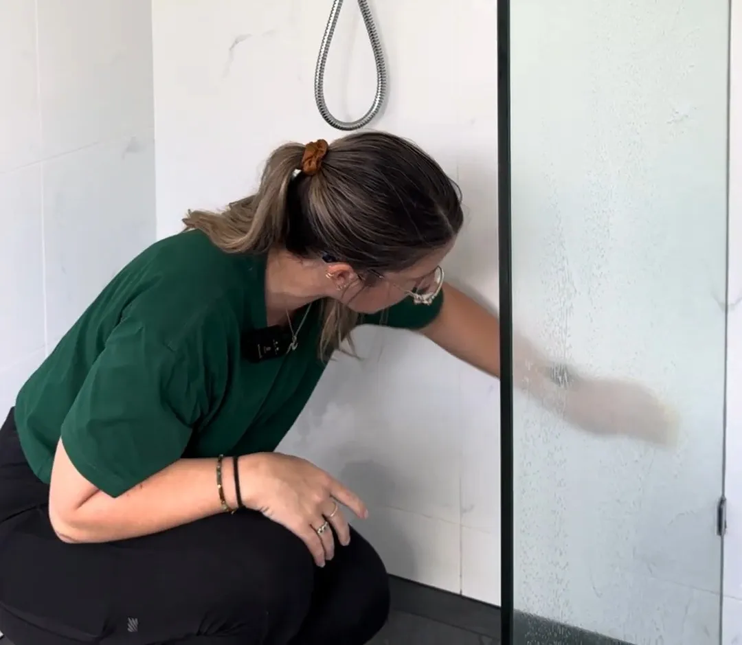 A TLB cleaner crouched in a walk-in shower, wiping down the tiles and glass