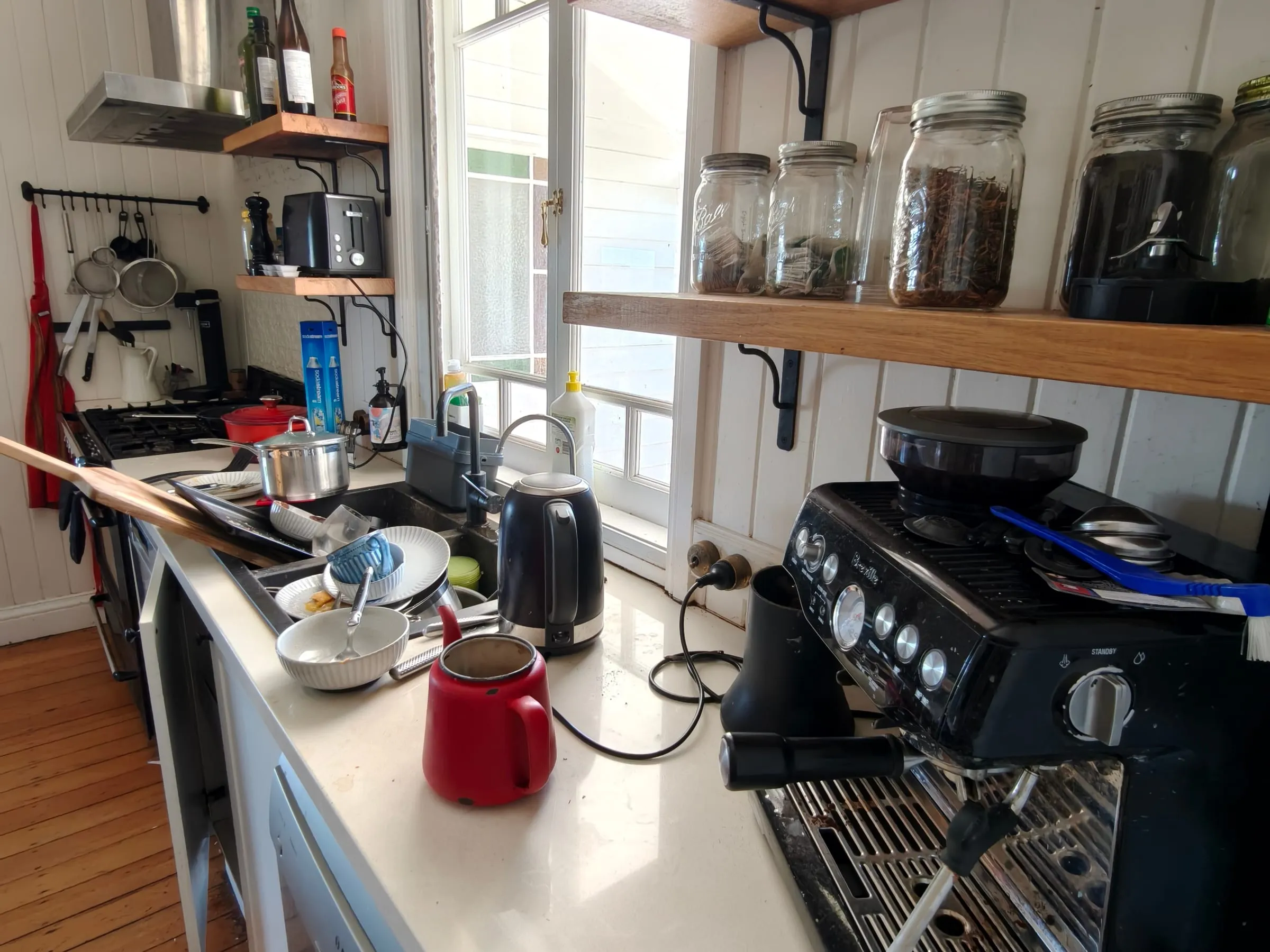 A kitchen benchtop before a TLB deep clean, with dishes stacked in and beside the sink and pots left on the stove