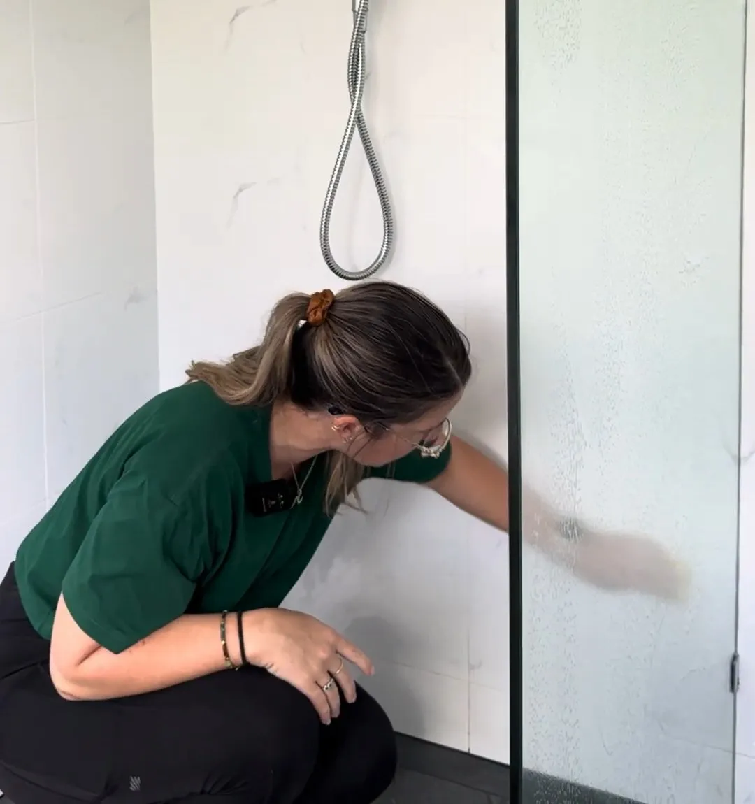 A TLB cleaner crouched in a walk-in shower, wiping down the tiles and glass