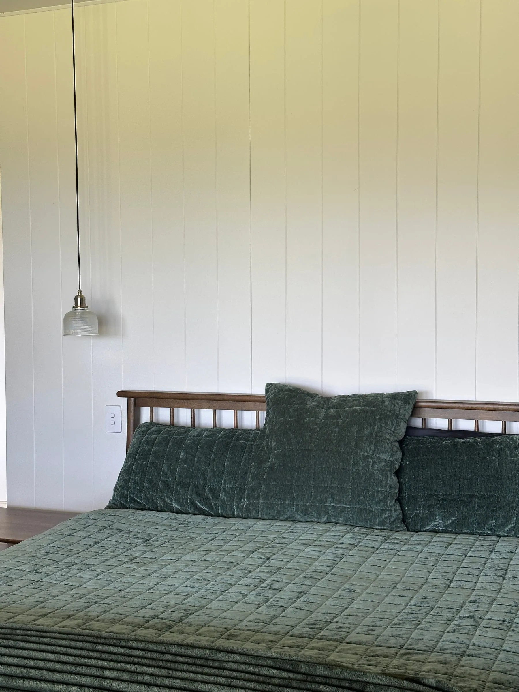 A freshly made bed with green quilted linen against a white panelled wall